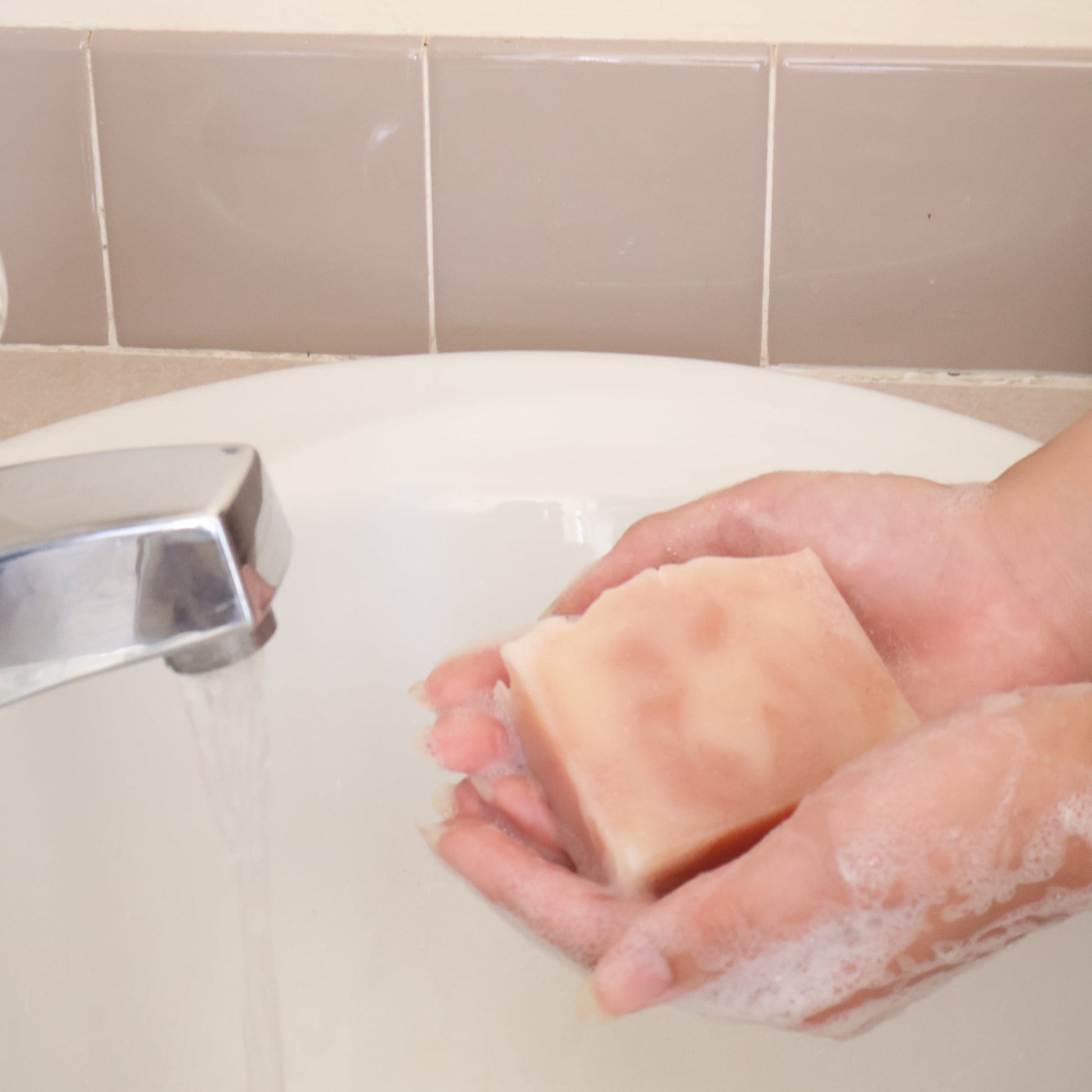 Hand holding a bar of soap over a sink with tiled walls in the background